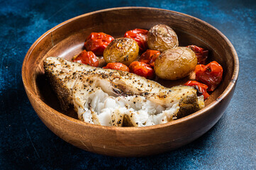 Baked halibut fish with roasted tomato and potato in wooden plate. Blue background. Top view. Copy...