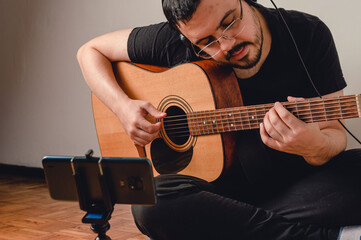 Obraz premium latin young man at home sitting on the floor playing guitar broadcasting live on his phone