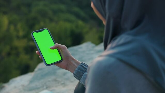 Woman in hijab on top of the hill in the evening uses smartphone with green screen on it. Focus on the cellphone. View under the shoulder