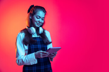 Portrait of young girl, student in shirt and checkered dress looking on tablet, posing isolated over pink studio background in neon light