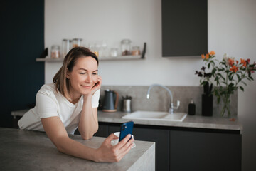 A young woman works remotely and chats on her smartphone in her kitchen at home.