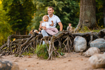 Naklejka na ściany i meble father and little cute blond daughter, together in the summer in nature, a little girl eats a lollipop, sitting on the roots of trees