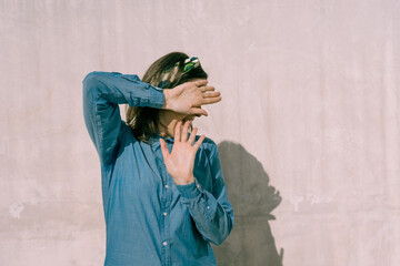 Brunette woman with blue shirt and floral print headband, turning and shielding her face with one hand, in front of a textured pinkish cream wall