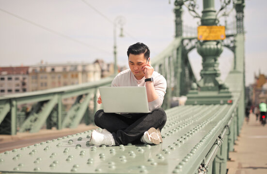 young man uses a computer and a smartphone sitting on a bridge