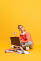 Portrait of young girl, student in sweater and glasses sitting on floor with thoughtful expression, studying isolated over yellow background