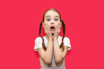 Fotobehang Girl child with pigtails on a red background with a surprised look holds on to her face. Shocked little girl with big eyes and open mouth. Child with a grimace of shock and surprise © halcon1