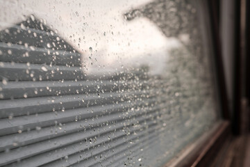 Raindrops on the glass of the window, which reflects the house and the cloudy sky, outdoors. 