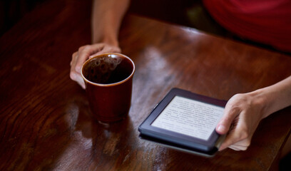 Woman reading e-book sitting on rural weathered wooden table with ceramic tea cup