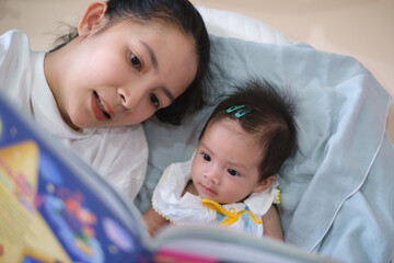 Smiling young Asian mother sit on the bed with little infant toddler child, happy biracial mom relax have fun read tale book with small baby girl at home, motherhood, childcare concept