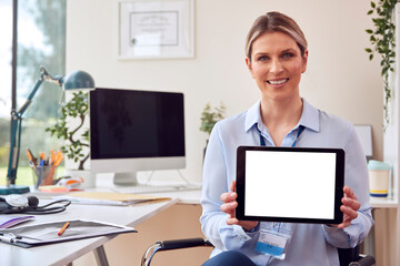 Portrait Of Smiling Female Doctor Or GP Sitting At Desk In Office Holding Digital Tablet