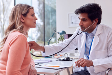 Male Doctor Or GP Wearing White Coat Examining Mature Woman Listening To Chest With Stethoscope