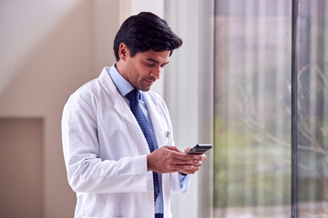Male Doctor Wearing White Coat Standing In Hospital Corridor Looking At Mobile Phone - Powered by Adobe