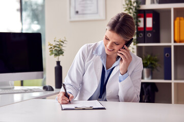 Smiling Female Doctor Or GP Wearing White Coat Sitting At Desk In Office Making Call On Mobile Phone