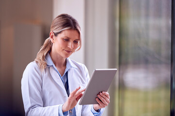 Female Doctor Wearing White Coat Standing In Hospital Corridor Looking At Digital Tablet