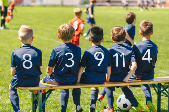 Football players wearing blue jersey shirts in the youth team sitting on a wooden bench. Multiracial group of kids in a school sports team. Boys in blue jersey shirts watching a tournament game