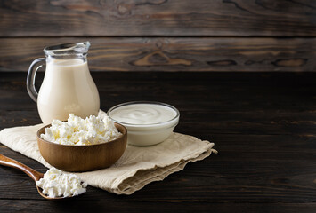 Milk cottage cheese in a wooden bowl on a light napkin. A jug of melted milk and sour cream in a transparent vase on a wooden table. Dark brown wooden background, space for text