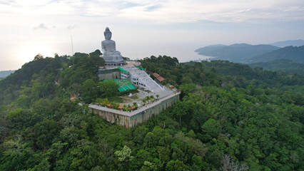 Drone view of the Big Buddha, Thailand. The Big Buddha is sitting on hill in the lotus position, meditating. The sun shines brightly through clouds. There's a jungle all around. A ray of sun on water