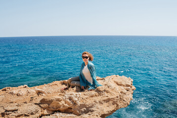 Young smiling girl sitting on yellow rock and looking at the camera on a background of blue sea on hot sunny day. Woman in t-shirt and scarf as protection from the sun. Tourist vacation. Copy space.