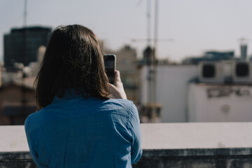 Woman with blue shirt taking photos with her phone on the roof of a building in Barcelona. Blurred building rooftops in the background. Film effect. Landscape.