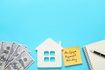 Note with words Mortgage Payment Holiday, house model, notebook and money on light blue background, flat lay. Space for text