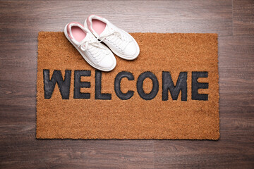 Stylish door mat with word Welcome and shoes on wooden floor, top view