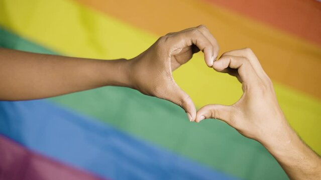 close up shot of hands making love or heart symbol in front of pride or rainbow flag - concept of gay couple love, solidarity and pride month celebration.