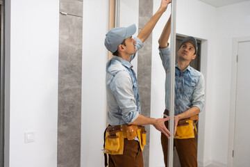 Employee furniture assembler in white t-shirt and jumpsuit checks wooden sliding door of modern wardrobe with mirror.