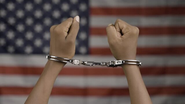 Close up shot of hanads rising with cuffs against us or american flag - concept of juneteenth, freedom and punishment.
