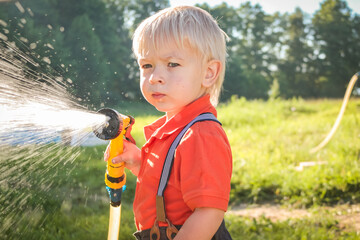 Cute little boy watering flowers in the garden. Young Child splashing Water from Garden Water Hose. Golden Hour Sunset. Very happy toddler with water sprinkler at summer - obrazy, fototapety, plakaty