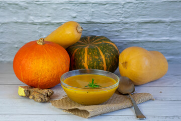 pumpkin soup in a glass bowl surrounded by different types of pumpkins and ginger on a wooden white...