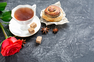 The concept of the celebration. A Cup of tea on a saucer, a red rose and a sweet bun on a dark background. Good morning. birthday. Space for text. Copy paste