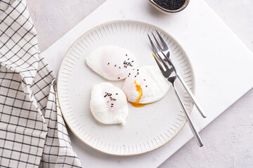 Three poached eggs with egg yolk dripping and black sesame seeds on a white plate on a marble board and silver forks