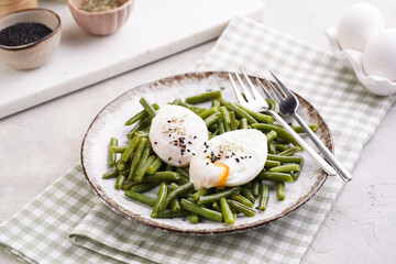 Soft-boiled poached eggs with black sesame seeds on a cooked green beans on a plate with brown rim on a green checkered kitchen towel