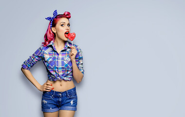 Portrait of beautiful woman licking heart shape lollipop dressed in pinup plaid shirt, isolated...