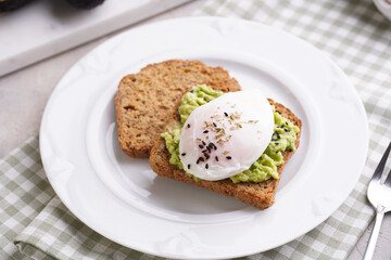 Slices of gluten-free sunflower seeds bread with mashed avocado, poached egg and sesame seeds on white plate on green checkered napkin