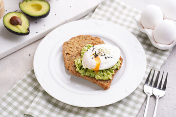Slices of gluten-free sunflower seeds bread with mashed avocado, poached egg and sesame seeds on white plate on green checkered napkin