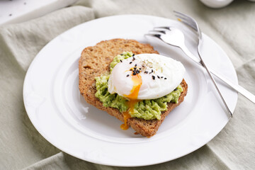 Slices of gluten-free sunflower seeds bread with mashed avocado, poached egg and sesame seeds on white plate on green checkered napkin