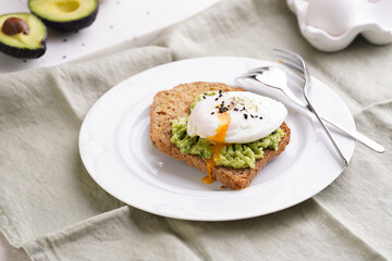 Slices of gluten-free sunflower seeds bread with mashed avocado, poached egg and sesame seeds on white plate on green checkered napkin