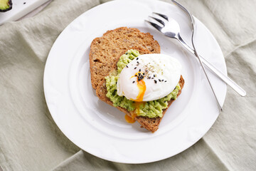 Slices of gluten-free sunflower seeds bread with mashed avocado, poached egg with egg yolk dripping and sesame seeds on white plate on green checkered napkin
