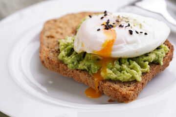 Slices of gluten-free sunflower seeds bread with mashed avocado, poached egg with egg yolk dripping and sesame seeds on white plate on green checkered napkin