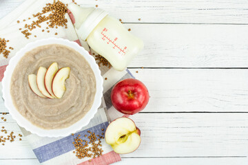 Fototapeta na wymiar Buckwheat porridge for a baby made of ground cereals with an apple in a white bowl, a bottle of milk on a white wooden background. Space for text. The first complementary food of a child.