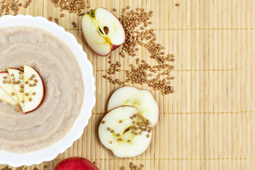 Buckwheat porridge for a baby made of ground cereals with a red apple in a white bowl on a brown background. Space for the text. The first complementary food of a child, baby nutrition.
