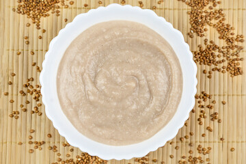 Buckwheat porridge for a baby from ground cereals in a white bowl on a brown bamboo background close-up. The first complementary food of a child, baby nutrition.