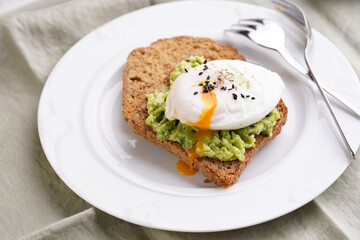 Slices of gluten-free sunflower seeds bread with mashed avocado, poached egg with egg yolk dripping and sesame seeds on white plate on green checkered napkin