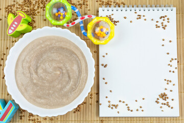 Buckwheat porridge for the baby from ground cereals in white bowl, rattle toys, a notebook with a clean page for writing on a bamboo background. Space for text. 