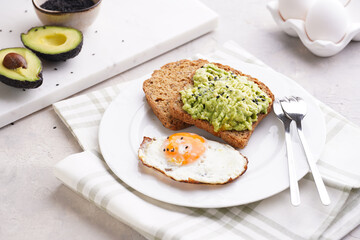 Slices of gluten-free sunflower seeds bread with mashed avocado, fried egg and sesame seeds on white plate on green checkered napkin