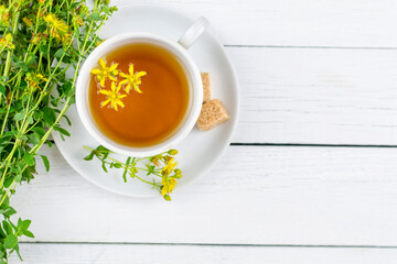 Herb and flowers of the medicinal plant Hypericum, Hypericaceae. A drink (infusion, decoction) of St. John's wort in a cup on a saucer on a white wooden background.