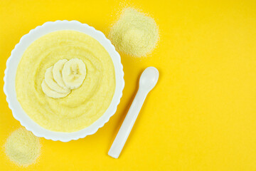 Corn porridge for a baby made of ground cereals in a white bowl with a banana, a spoon on a bright yellow background. The first complementary food of a child, baby nutrition. Space for the text.