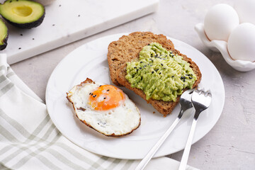 Slices of gluten-free sunflower seeds bread with mashed avocado, fried egg and sesame seeds on white plate on green checkered napkin
