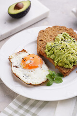 Slices of gluten-free sunflower seeds bread with mashed avocado, fried egg and sesame seeds on white plate on green checkered napkin